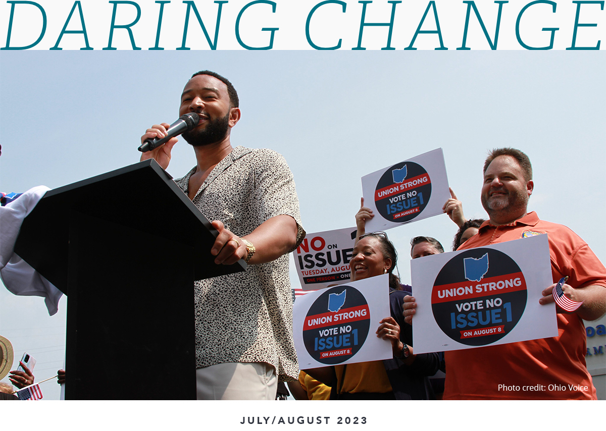John Legend standing at a podium holding a microphone with a multi-racial group of people behind him holding up signs that read ‘Union Strong. Vote Issue #1.’