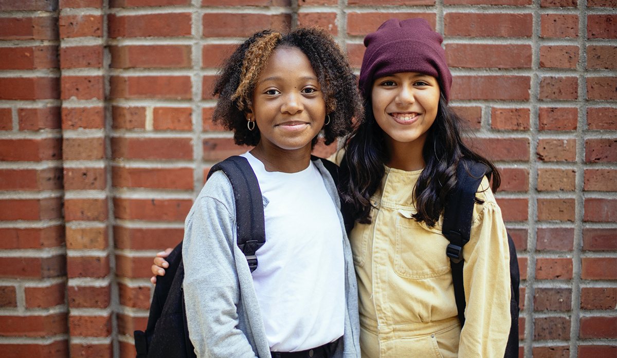 A young Black girl and a young Latine girl wearing school backpacks, smiling and posing for the camera together against a brick wall.