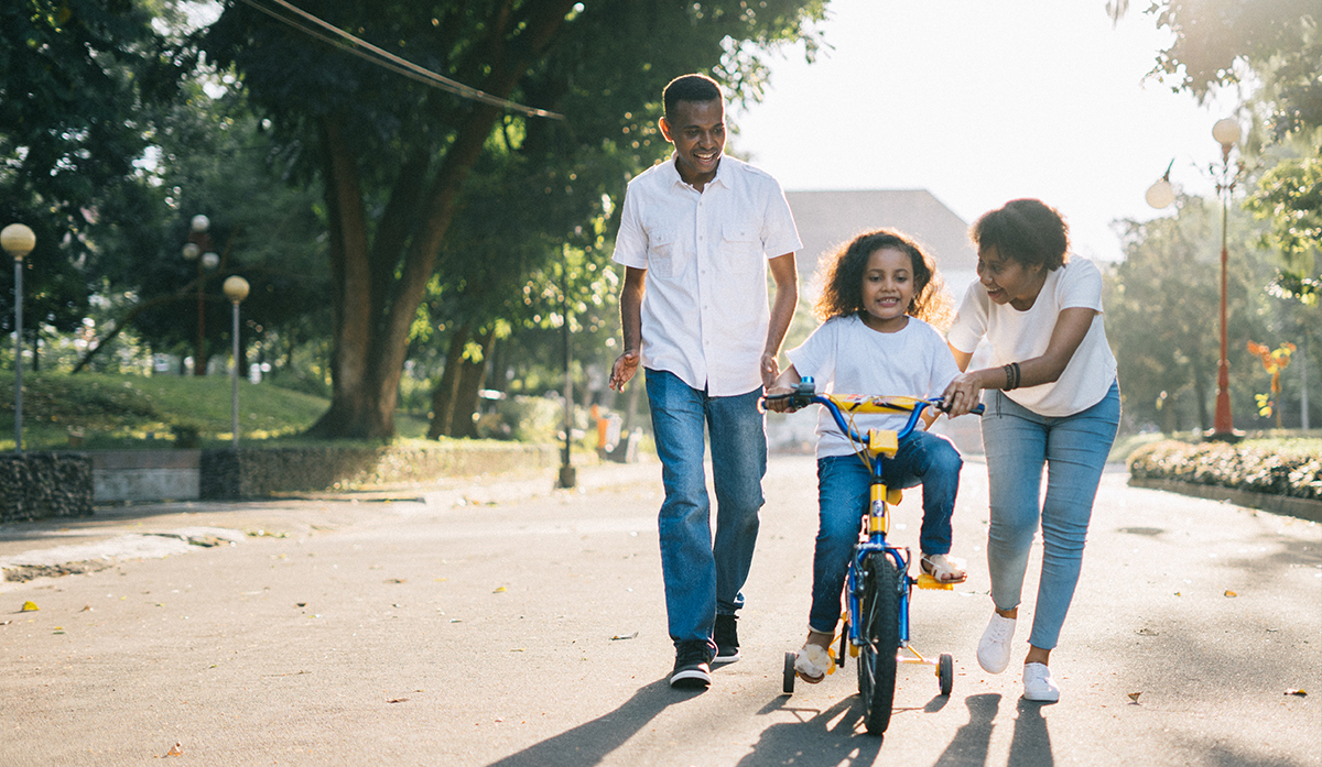 A young Black girl learning how to ride a bike with her mom and dad.